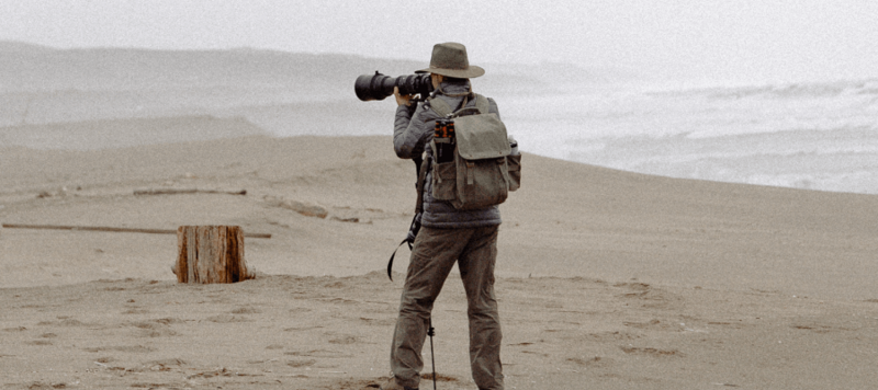 Photographer with telephoto lens and backpack standing on misty beach, shooting toward ocean.