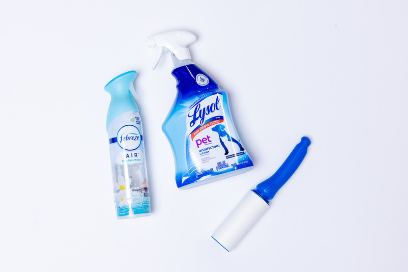 Three cleaning products arranged on a white surface: an air freshener spray, a pet-specific disinfectant spray, and a lint roller with blue handle.
