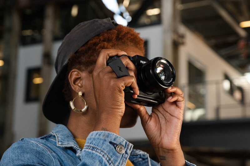 Photographer holding vintage rangefinder camera up to face, wearing denim shirt and dark cap.