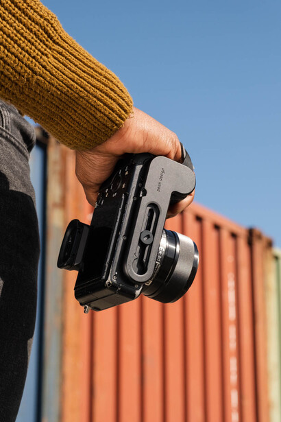 Photographer holding a compact digital camera against a corrugated metal backdrop in bright sunlight.