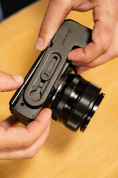 Hands holding a compact rangefinder camera with attached lens against yellow background.