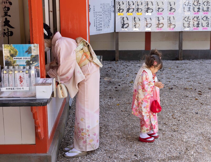 Woman in pink kimono viewed from behind standing near a small child in floral kimono at a Japanese temple.
