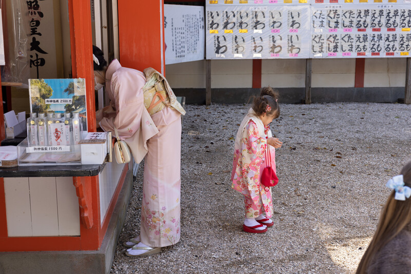 Child in pink floral kimono standing in temple courtyard near bulletin boards and shop window.