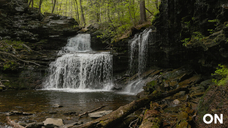 Multi-tiered waterfall cascading into a pool surrounded by moss-covered rocks and dense forest.