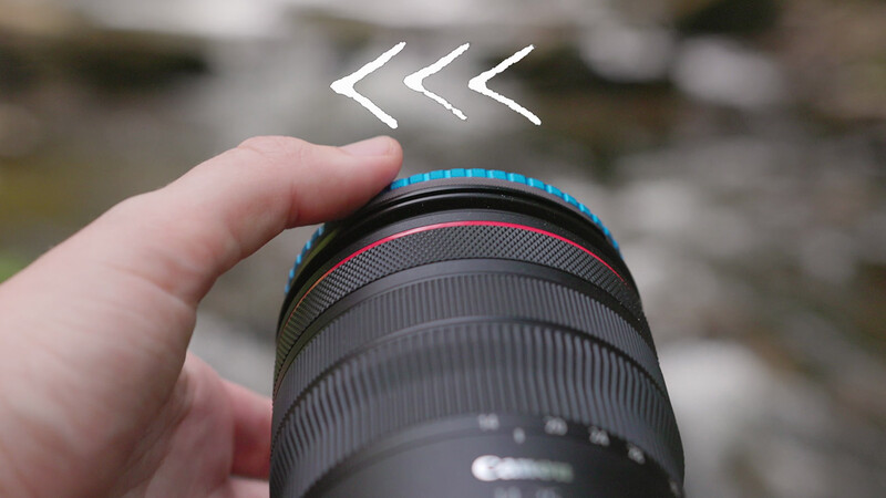 Close-up of a camera lens with blue focus ring and red accent line held in a hand against blurred background.