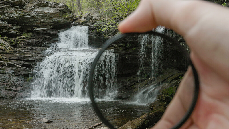 Circular polarizing filter held up to a cascading waterfall surrounded by moss-covered rocks and forest vegetation.
