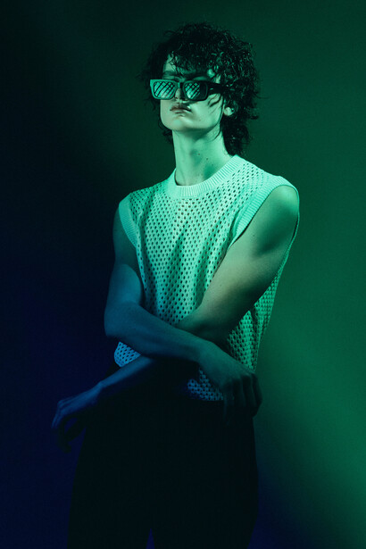 Person wearing sunglasses and textured sleeveless top posing against gradient teal and blue background.