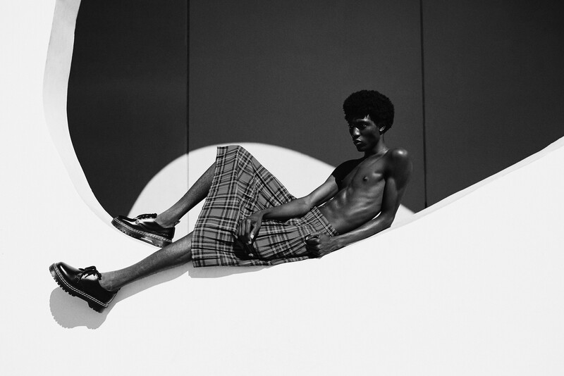 Shirtless man reclining against curved geometric shapes, wearing plaid shorts and sunglasses.