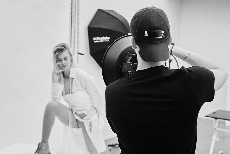 Photographer with camera and lighting equipment shooting a model in a white shirt during studio session.