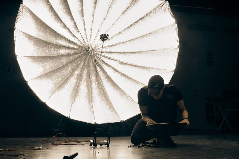 Photographer crouching beside large parabolic reflector producing bright directional light against dark background.