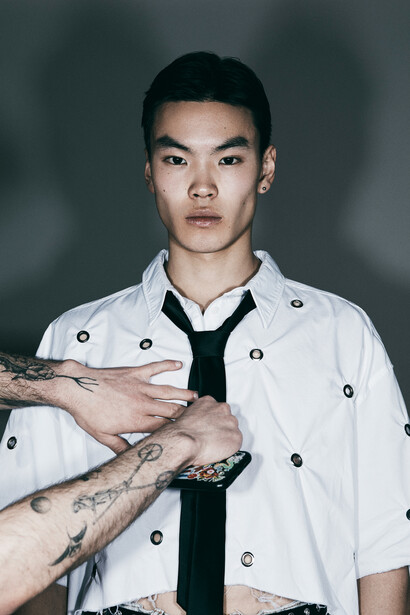 Portrait of a young man with visible arm tattoos wearing a white button-up shirt and black tie against a neutral gray background.