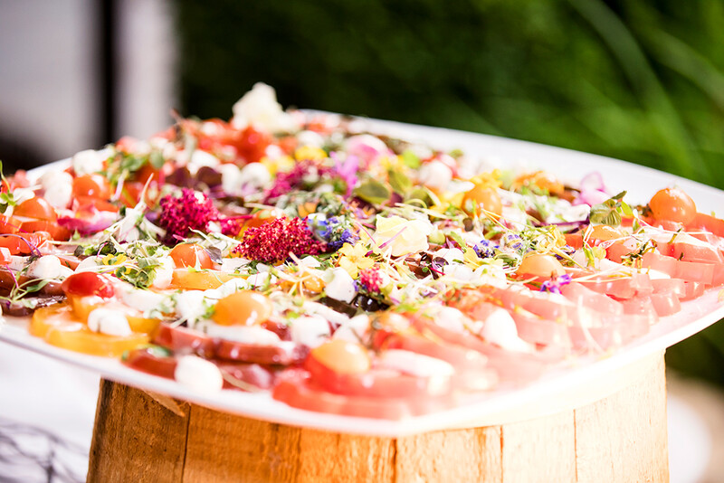Colorful appetizer platter with cured meats, fresh vegetables, and edible flowers arranged on a wooden base.