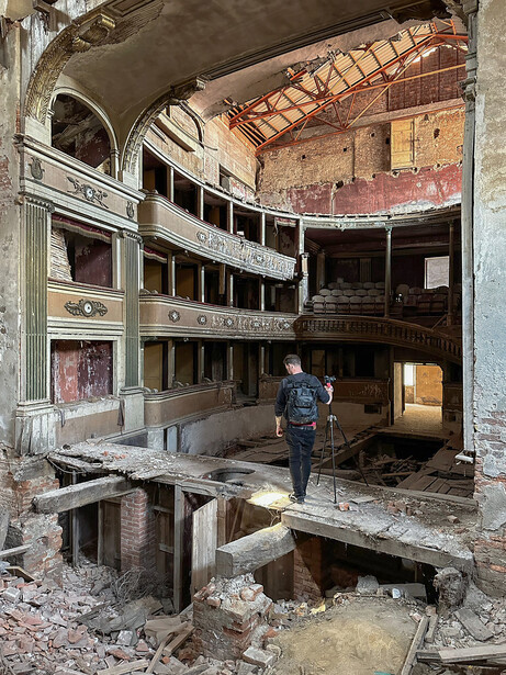 Person standing in the debris-filled interior of an abandoned multi-story theater with exposed structural elements and deteriorating architecture.