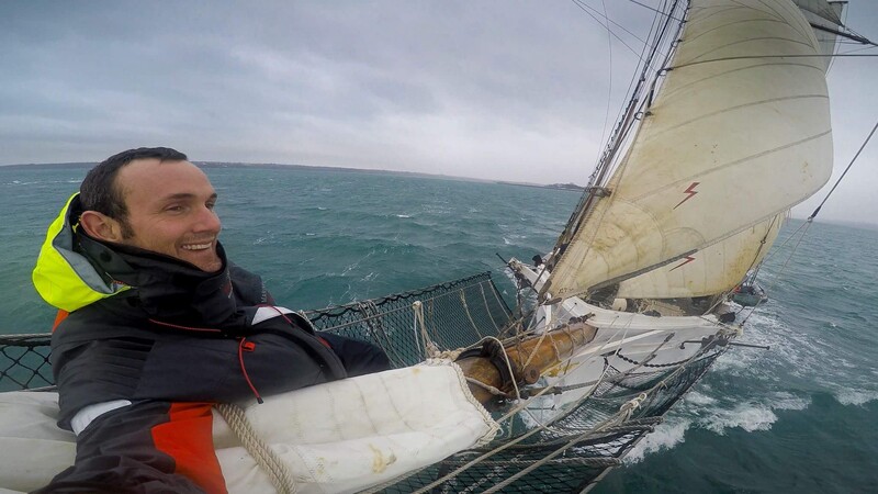 Sailor smiling at camera while holding rope on tilting sailboat with full sails deployed.