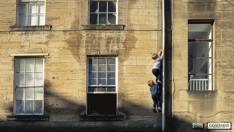 Two people climbing on the exterior of a golden-stone building facade alongside a dark doorway.