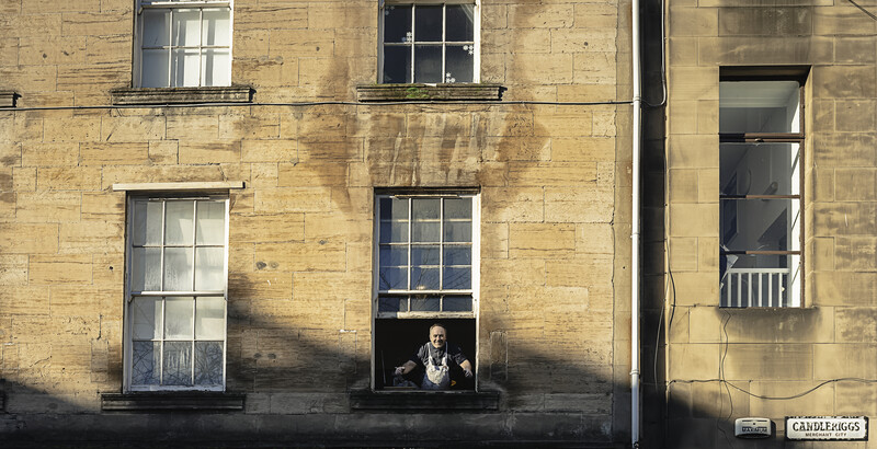Person looking out from a window in a weathered stone building facade with symmetrical windows and strong shadow patterns.