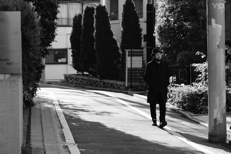 Solitary figure in dark clothing walking through a modern courtyard with strong diagonal sunlight and architectural shadows.