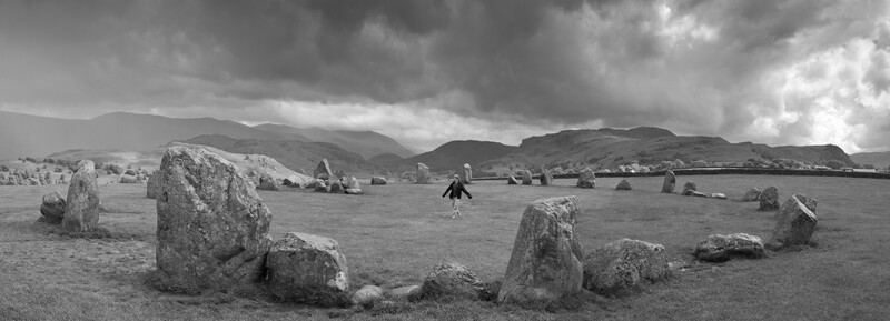 Stone circle with scattered boulders on grassy landscape under dramatic cloudy sky.