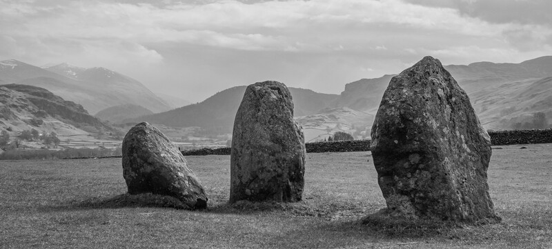 Three standing stones in a field with mountains and a river valley in the background.
