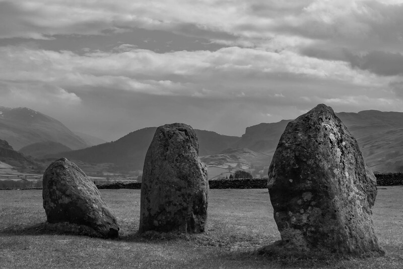 Three weathered standing stones arranged in a grassy field with mountains and dramatic clouds beyond.