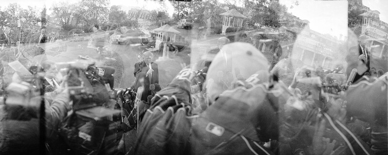 Blurred black and white photograph of a crowded outdoor gathering with residential houses and trees in background.