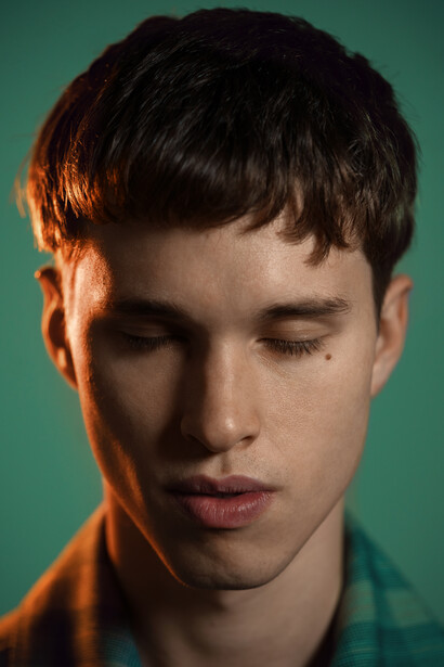 Young man with dark hair and closed eyes against teal background, lit by warm side light.