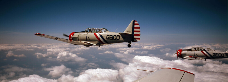 Two GEICO-branded aerobatic aircraft flying in formation above a cloud layer against a clear blue sky.