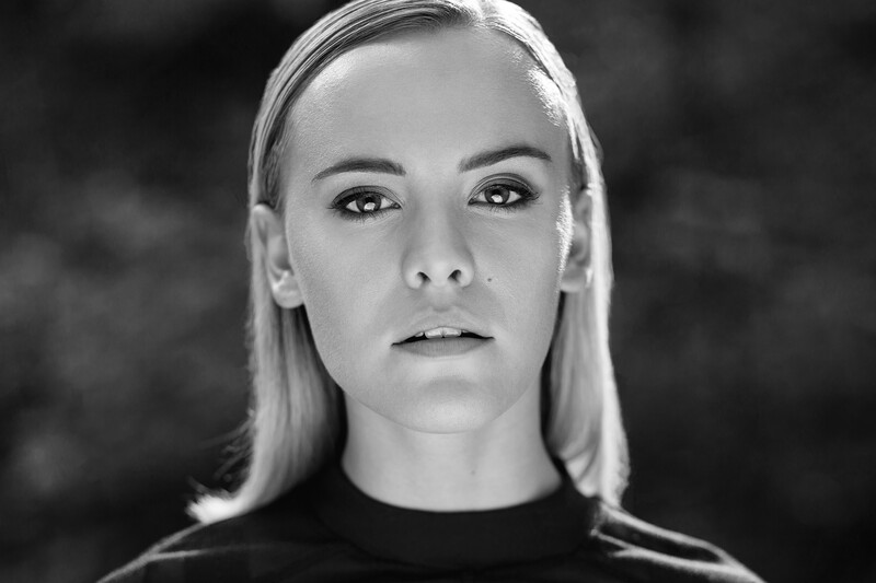 Black and white portrait of a woman with straight blonde hair and direct gaze against a dark background.