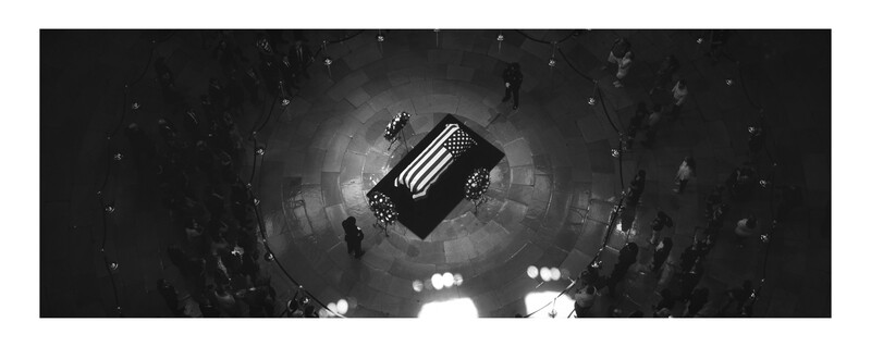Overhead view of a flag-draped casket surrounded by mourners in a circular formation under dramatic lighting.