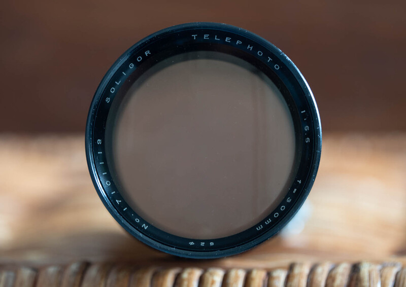 Vintage telephoto lens viewed front-on, showing blue barrel and glass optics against blurred background.