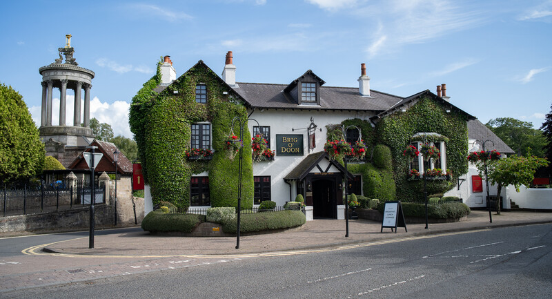 Ivy-covered historic pub with white and dark timber framing, ornate chimneys, and decorative planters beside a cylindrical stone monument.