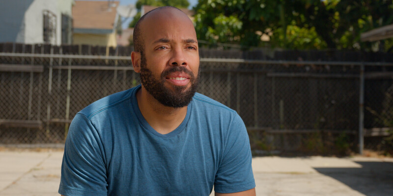 Man with beard wearing blue v-neck t-shirt outdoors in bright daylight.