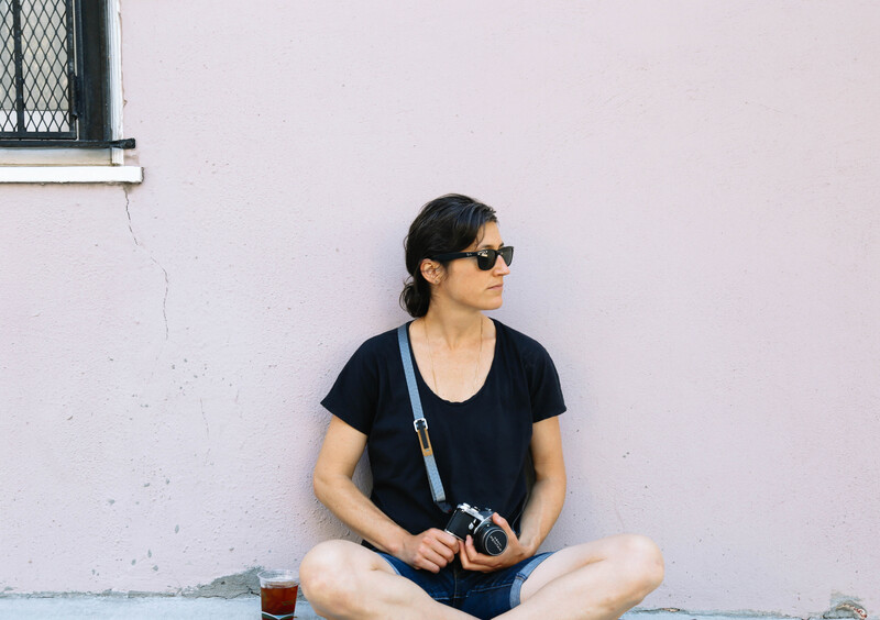 Woman in black t-shirt sitting cross-legged against white wall, holding vintage camera.