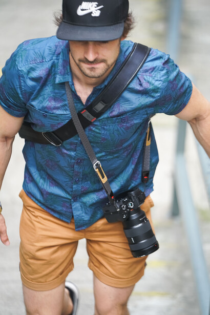 Male photographer wearing denim shirt and khaki shorts with camera bag slung across chest.