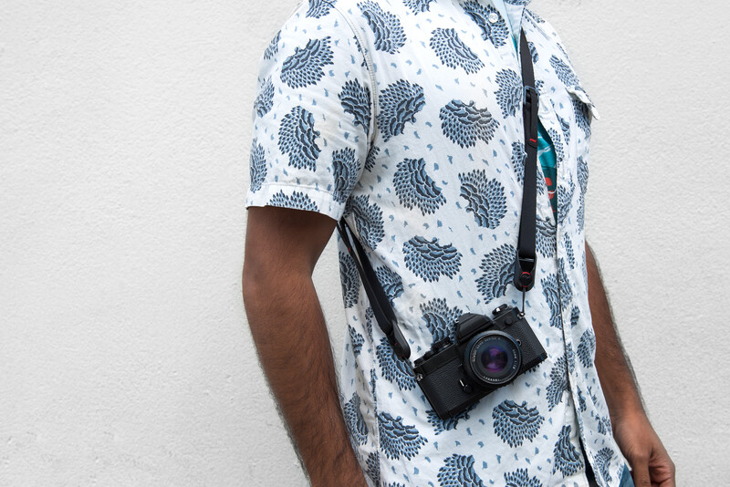 Person wearing a leaf-patterned shirt holding a compact camera with a shoulder strap.