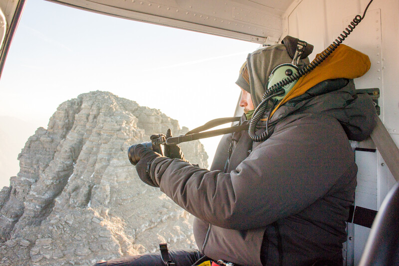 Photographer shooting from helicopter with telephoto lens aimed at rocky mountain peak.