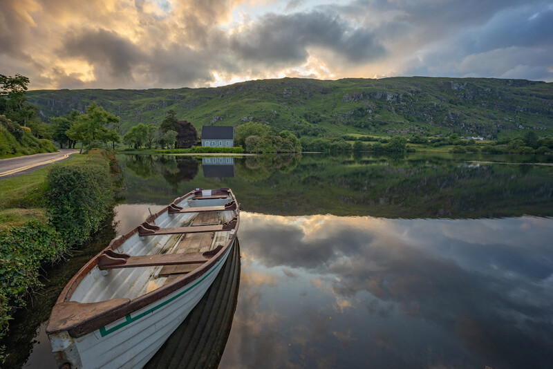 Wooden rowboat moored on still water with forested hills and dramatic clouds reflected in the glassy surface.