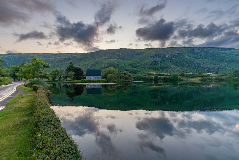 Calm river reflecting dramatic clouds with green hillsides and a small building along the grassy bank.