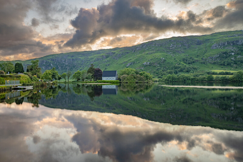 A stillwater lake reflects dramatic clouds and a verdant hillside with scattered trees and buildings.