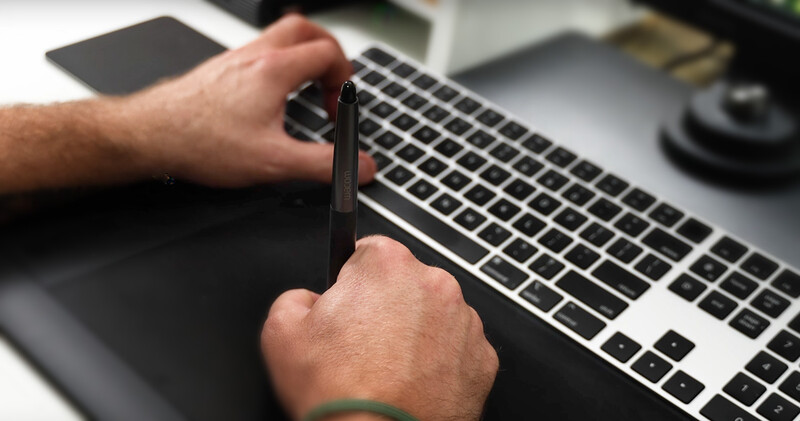 Hands holding a stylus pen above a laptop keyboard in a studio workspace.