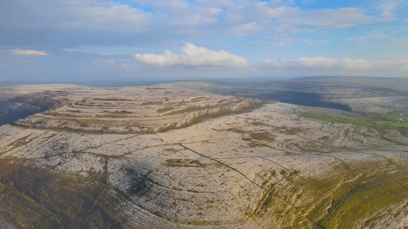 Aerial view of rolling limestone hills with agricultural fields and patches of woodland beneath partly cloudy sky.
