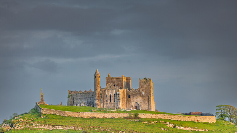 Medieval stone castle with multiple towers set on a green hilltop beneath dramatic cloudy sky.