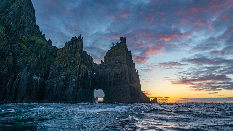 Dramatic rocky sea stacks silhouetted against a colorful sunset sky with golden and pink clouds.
