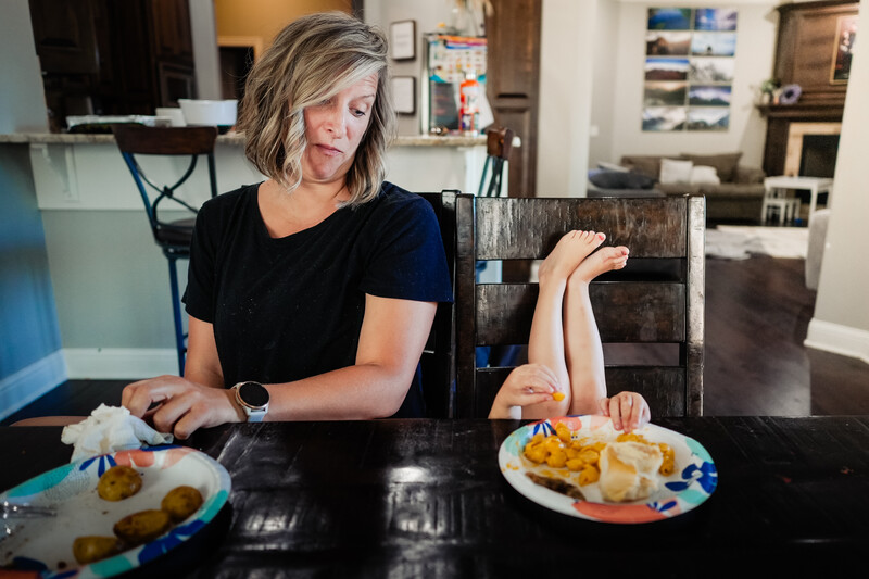 Woman at a dining table with plates of food, resting her arm on a wooden chair.