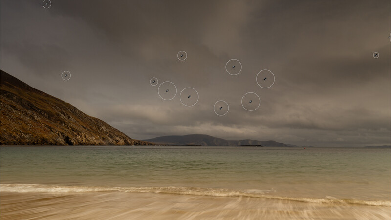 Coastal landscape with sandy beach, turquoise water, and rocky headland under stormy sky with floating circles.