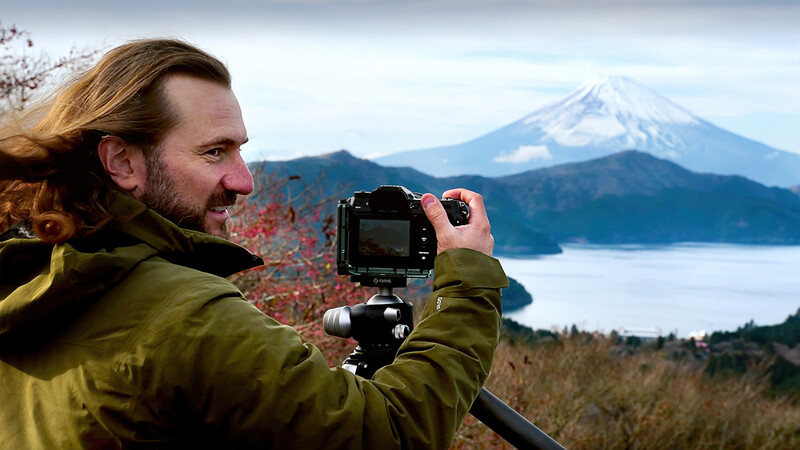 Photographer operating a camera on a tripod while composing a landscape shot of a snow-capped volcanic mountain across a lake.