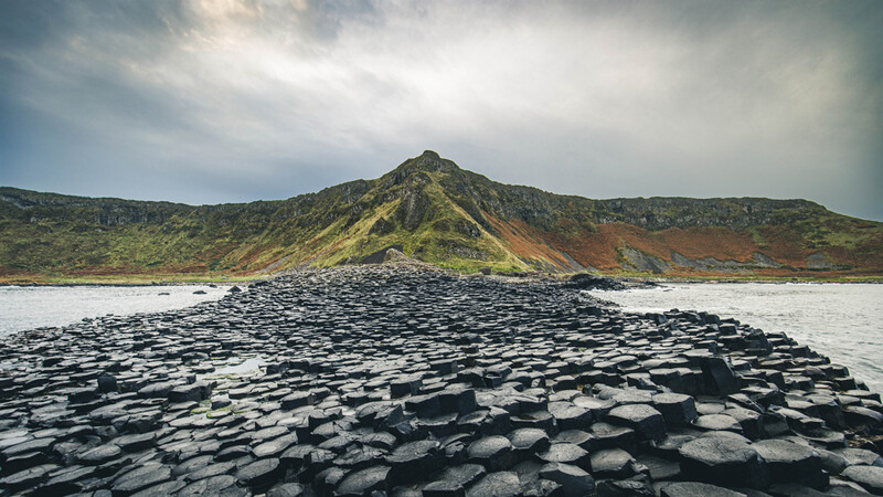 Black hexagonal basalt columns extend into shallow water with a moss-covered volcanic mountain in the background.
