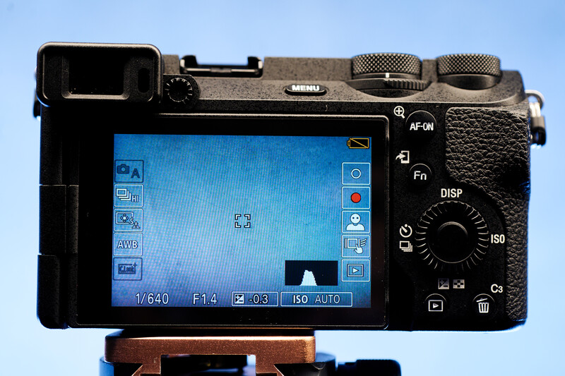 Screenshot of a mirrorless camera's rear LCD display showing menu options and shooting information against a blue sky background.