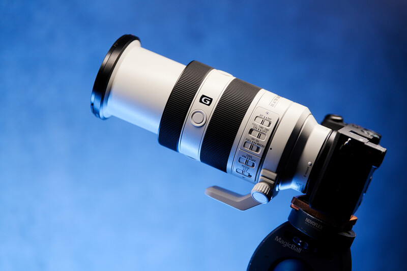 White telephoto lens with control rings mounted on a black gimbal head against a blue background.