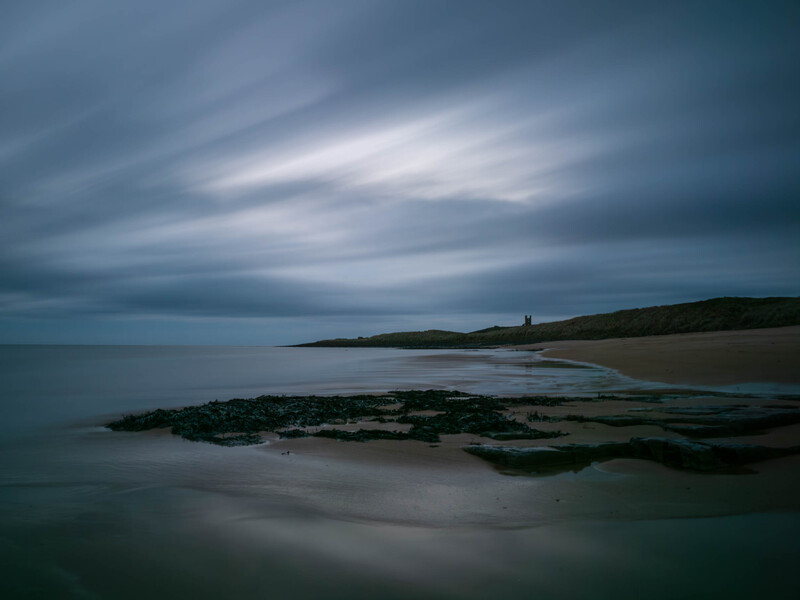 Moody coastal landscape with rocky foreground and distant headland under dramatic clouded sky.